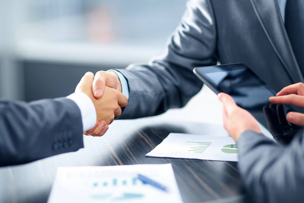 People in suits shaking hands across a table with another using a tablet 